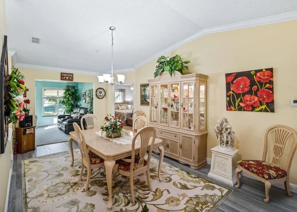 Inviting dining area featuring wood-look vinyl flooring, crown molding, and a vaulted ceiling with a chandelier. The open layout flows into a sunlit sitting area, creating a welcoming space for everyday meals and entertaining.
