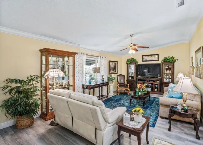 Bright and inviting living room featuring wood-look vinyl flooring, crown molding, and a ceiling fan. A large window welcomes natural light, while soft neutral walls create a comfortable setting for relaxing or entertaining.