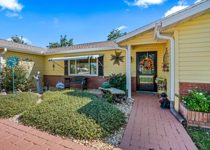 Welcoming covered entry with a brick-paver walkway, cheerful yellow siding, and classic brick accents. Landscaped garden beds, a cozy bench beneath a striped window awning, and decorative touches create a charming first impression.