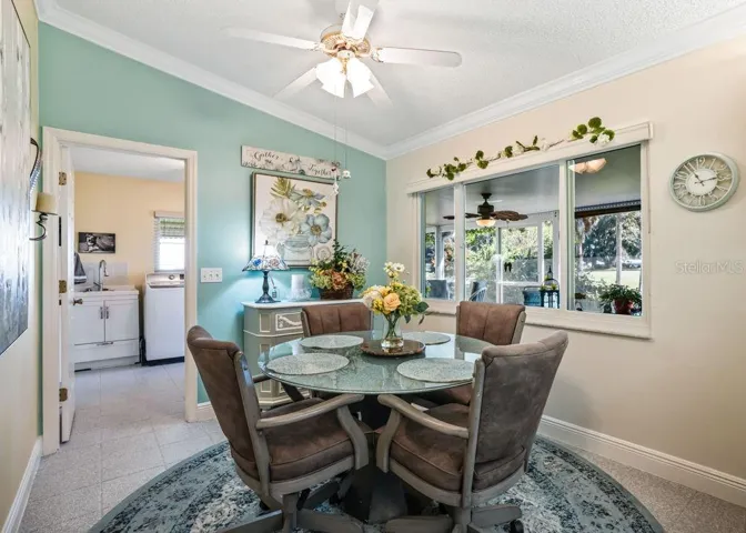 Charming breakfast nook featuring a soft aqua accent wall, crown molding, and a ceiling fan. A large interior window overlooks the enclosed lanai and greenery beyond, while the adjacent laundry room adds everyday convenience.
