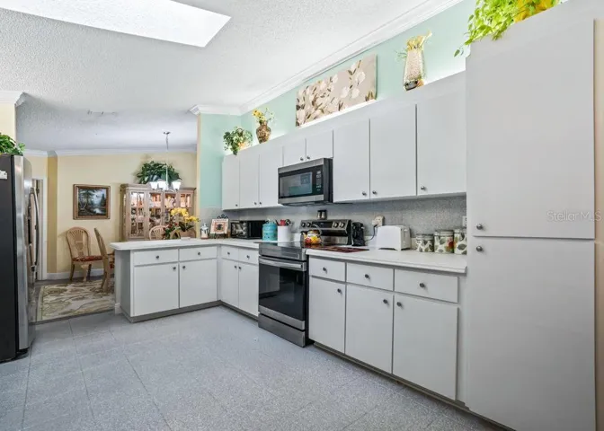 Inviting kitchen with white cabinetry, generous counter space, a tall pantry cabinet, and stainless steel appliances. A skylight brightens the space, while aqua accents and crown molding add charm. The adjoining dining area offers a convenient flow for meals and entertaining.