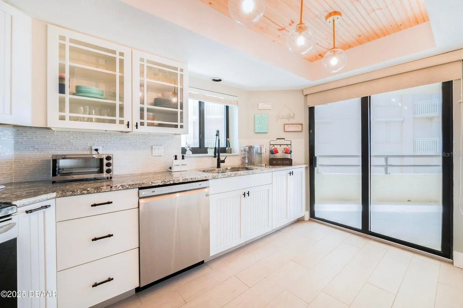 Beautifully appointed kitchen featuring white cabinetry, granite countertops, a stylish tile backsplash, and glass-front upper cabinets. A wood-accented tray ceiling with globe pendant lighting adds warmth and architectural character.