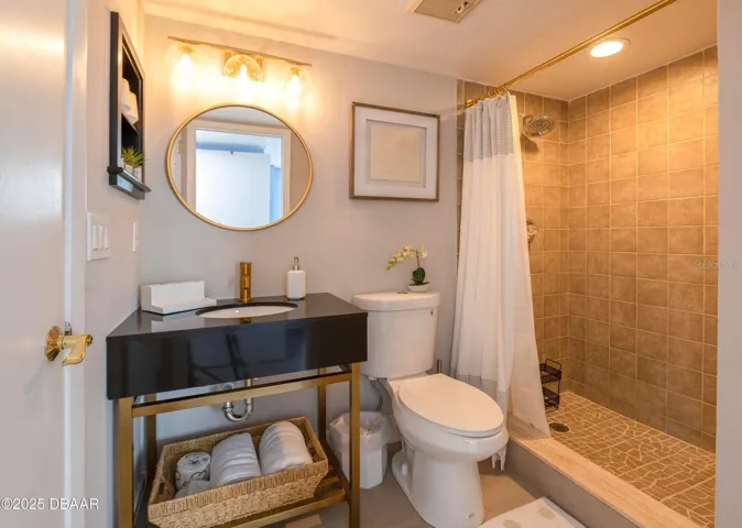 This stylish guest bathroom features a modern black vanity with gold-tone accents, a round mirror, updated lighting, and a tiled walk-in shower with a rainfall showerhead.