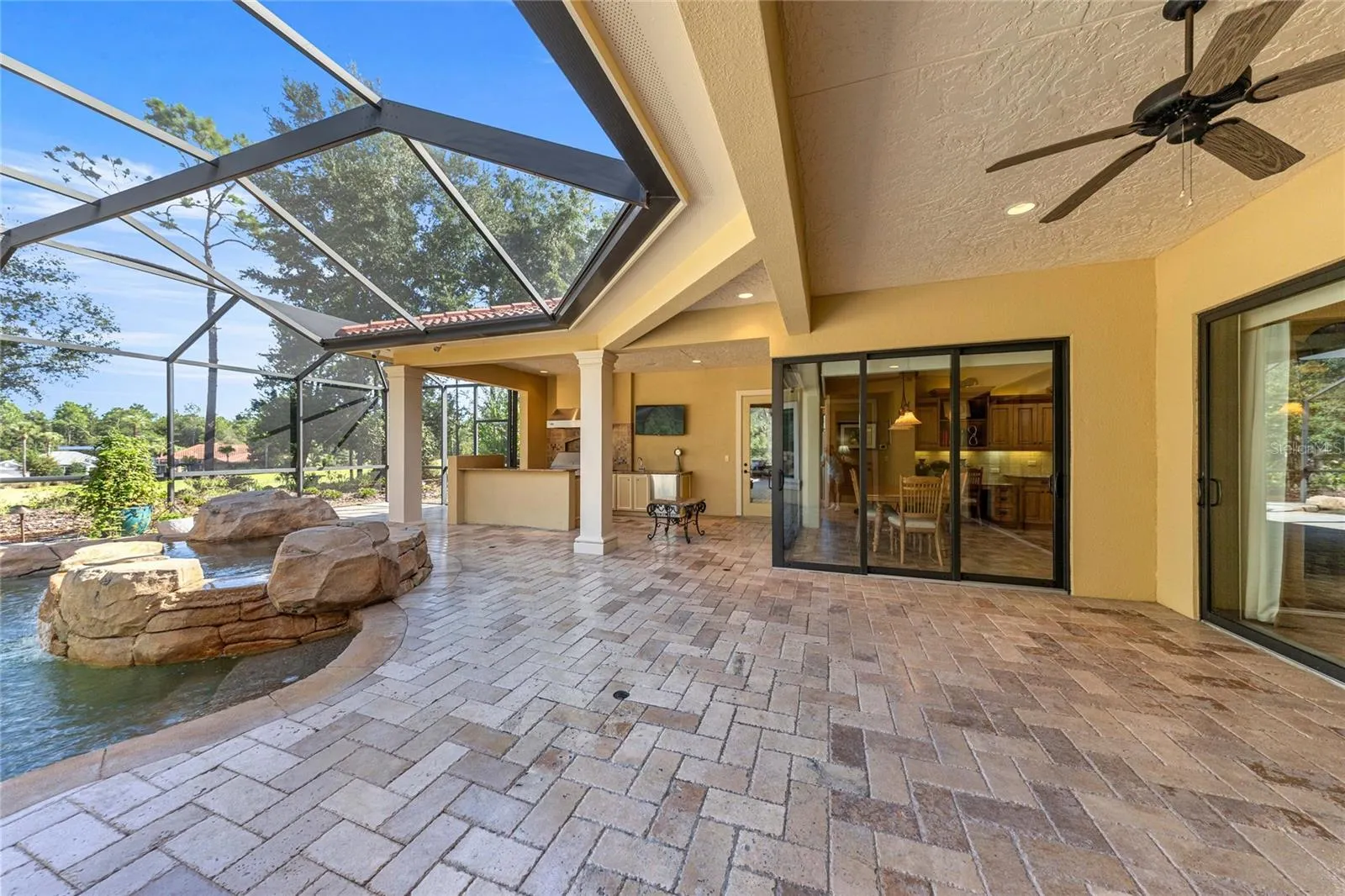 Expansive covered lanai with brick pavers, ceiling fan, and views of the custom pool and spa.