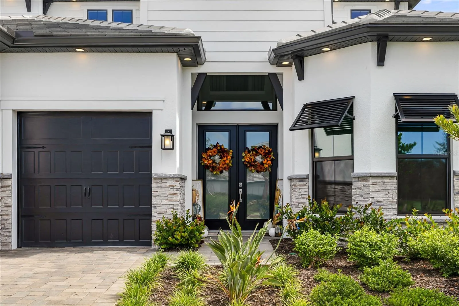 Elegant two-story residence featuring a striking modern coastal exterior, lush tropical landscaping, and an inviting paver driveway leading to a spacious three-car garage.