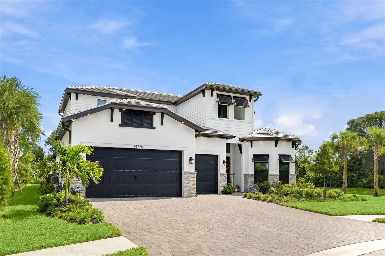 Elegant two-story residence featuring a striking modern coastal exterior, lush tropical landscaping, and an inviting paver driveway leading to a spacious three-car garage.