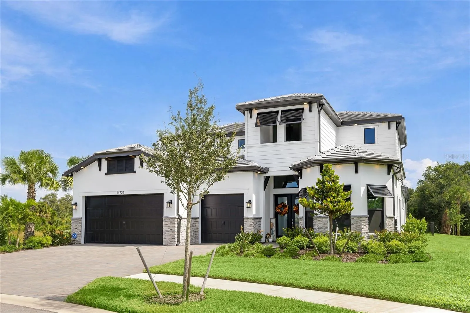 Elegant two-story residence featuring a striking modern coastal exterior, lush tropical landscaping, and an inviting paver driveway leading to a spacious three-car garage.