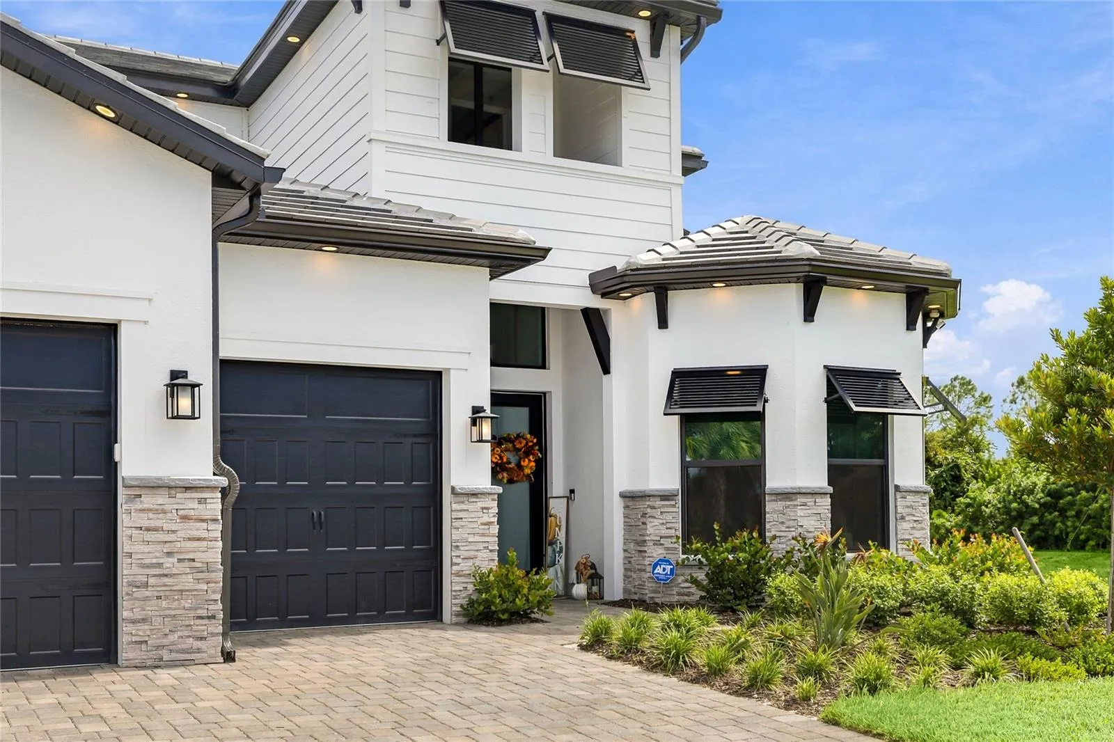 Elegant two-story residence featuring a striking modern coastal exterior, lush tropical landscaping, and an inviting paver driveway leading to a spacious three-car garage.