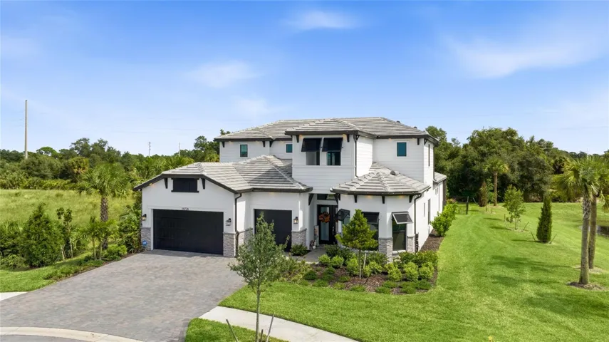 Elegant two-story residence featuring a striking modern coastal exterior, lush tropical landscaping, and an inviting paver driveway leading to a spacious three-car garage.