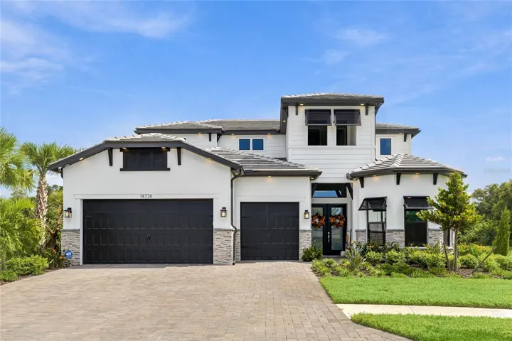 Elegant two-story residence featuring a striking modern coastal exterior, lush tropical landscaping, and an inviting paver driveway leading to a spacious three-car garage.