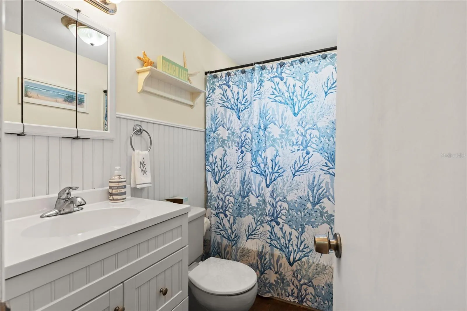 Coastal charm continues into this full bathroom, featuring a white vanity with decorative beadboard-style detailing, a mirrored medicine cabinet, and a tub/shower combination. White wainscoting and cheerful blue accents complete the beach-inspired look.