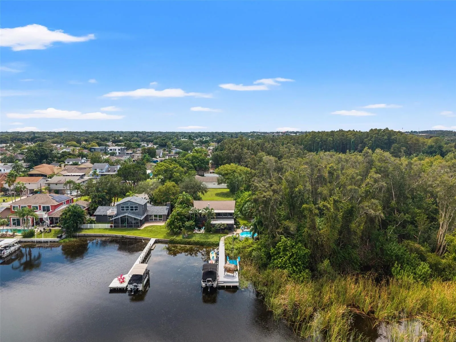 Aerial photography highlights the property's setting alongside a heavily wooded natural area, with neighboring waterfront homes and private docks lining the shoreline. The combination of mature trees, tropical landscaping, and water access creates an appealing Florida lifestyle.