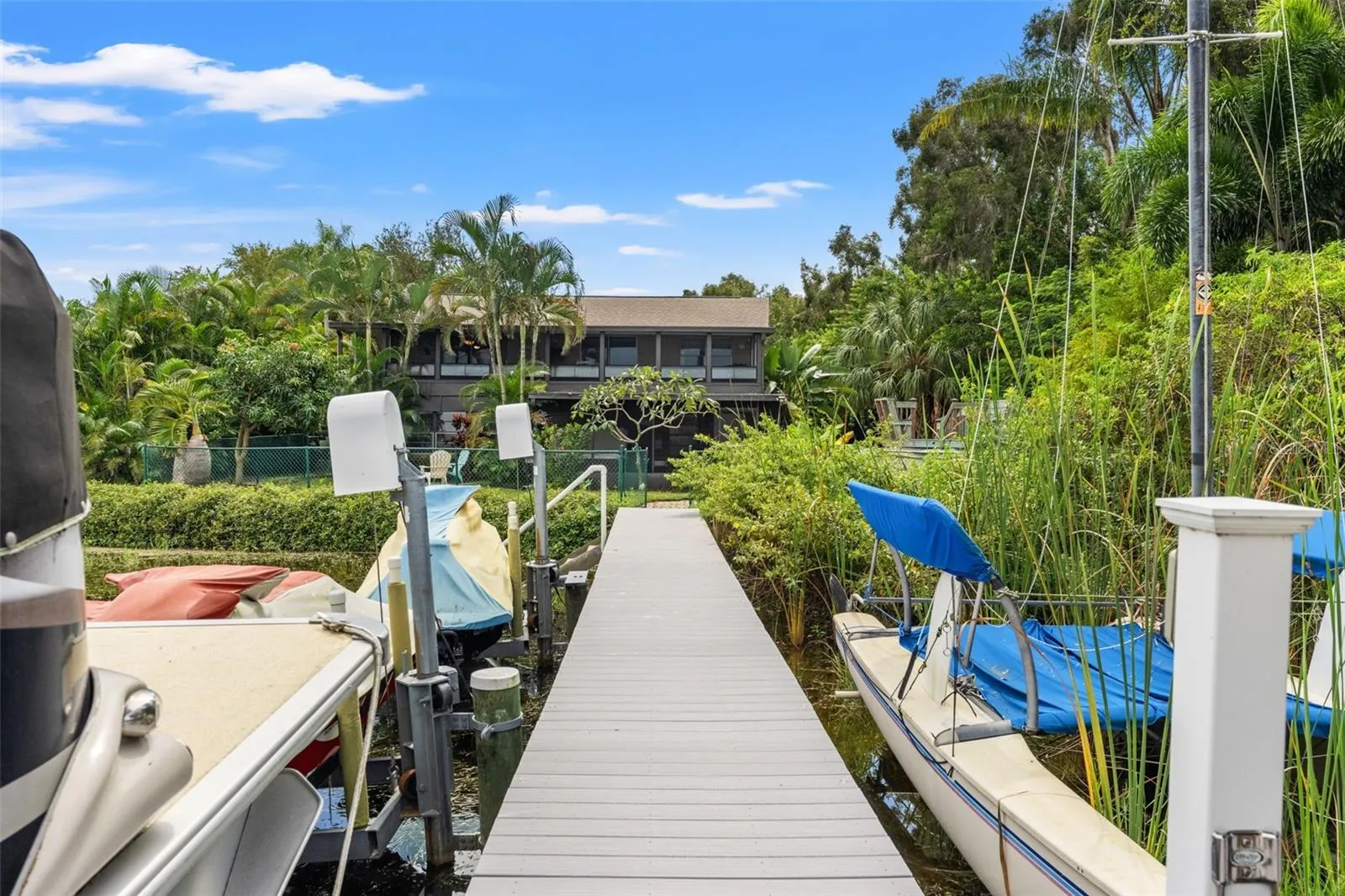 A view from the dock looking back toward the home showcases its elevated design, tropical landscaping, and waterfront setting. The long dock connects the backyard to the lake, with boat and watercraft lifts providing convenient access for outdoor recreation.