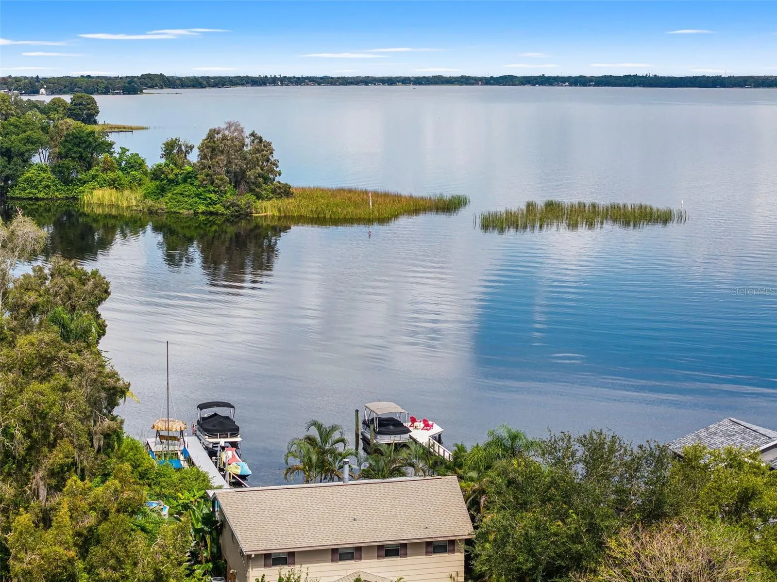 An elevated perspective highlights the property's dock, pontoon boat lift, and thatched-roof seating area. Beyond the shoreline, expansive open water stretches toward the distant tree line, offering an inviting setting for boating, fishing, and enjoying the outdoors.