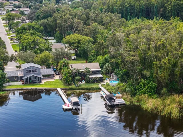 This aerial photograph brings together several of the property's standout features: an elevated home, above-ground backyard pool, private dock with pontoon boat lift, and shaded thatched-roof swing. The adjoining wooded area creates a natural backdrop, making the waterfront setting especially appealing.