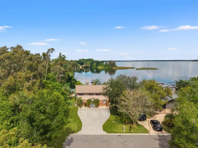 This aerial perspective captures the home's setting along a quiet residential street, surrounded by mature trees with the expansive lake stretching out behind it. The elevated home enjoys a generous driveway, lush greenery, and a waterfront location that makes boating and outdoor living part of everyday life.