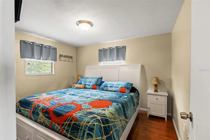 A comfortable bedroom with warm wood flooring, neutral walls, and two windows providing natural light. The room accommodates a generously sized bed with nightstands, while its coastal-inspired décor complements the home's relaxed waterfront setting.