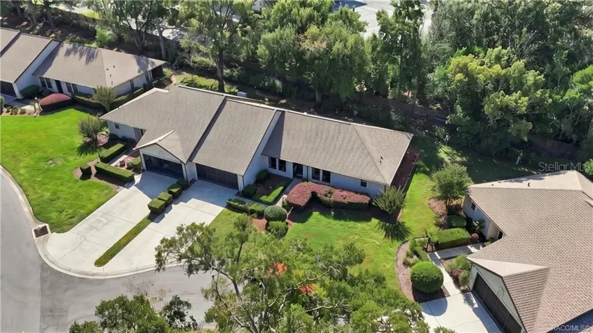 Aerial perspective showcasing the home's roofline, beautifully maintained grounds, surrounding residential properties, and wooded backdrop.