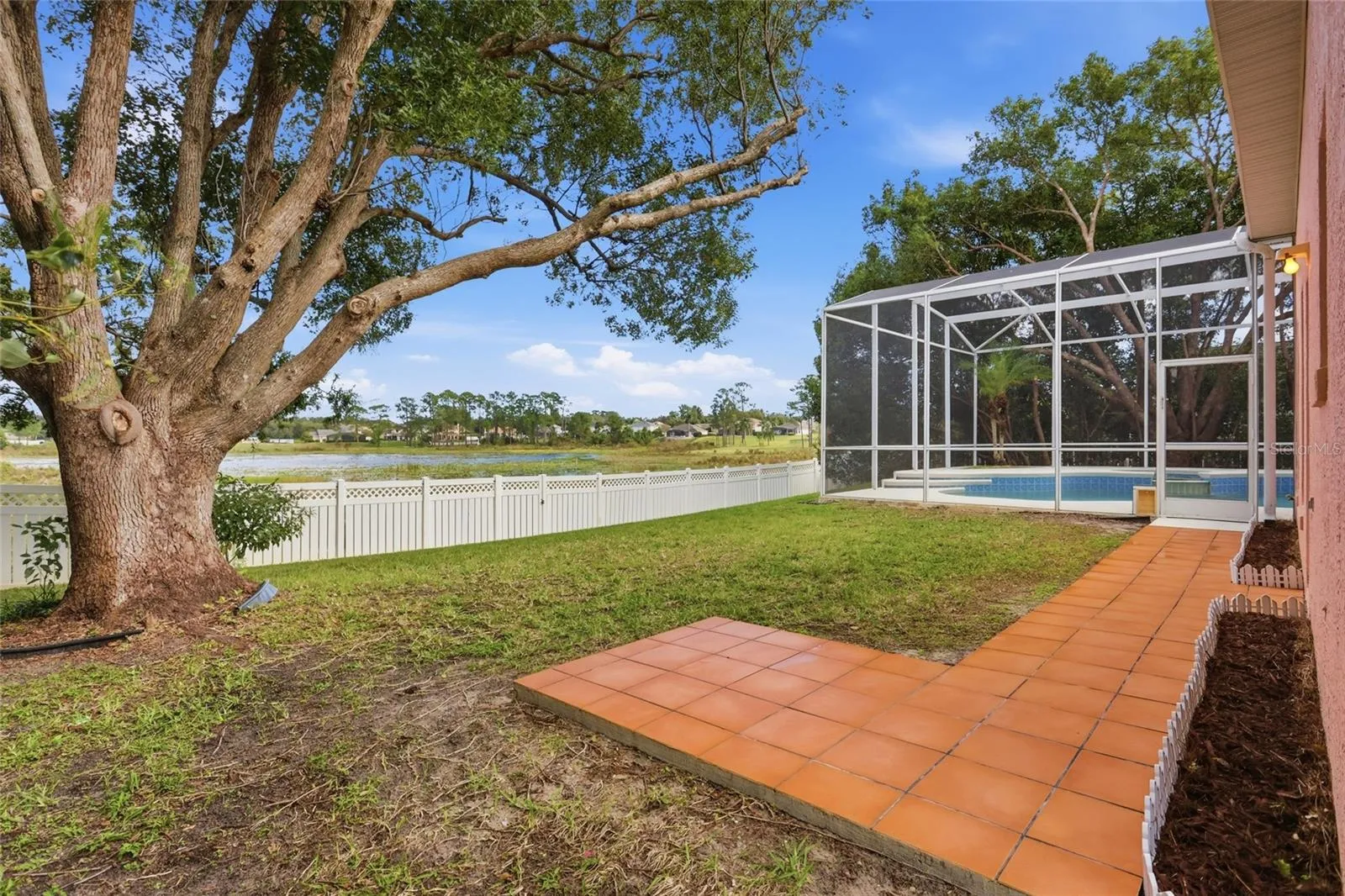 Mature trees shade the backyard beside the pool enclosure, with a paved path and lakeviews beyond the fence.