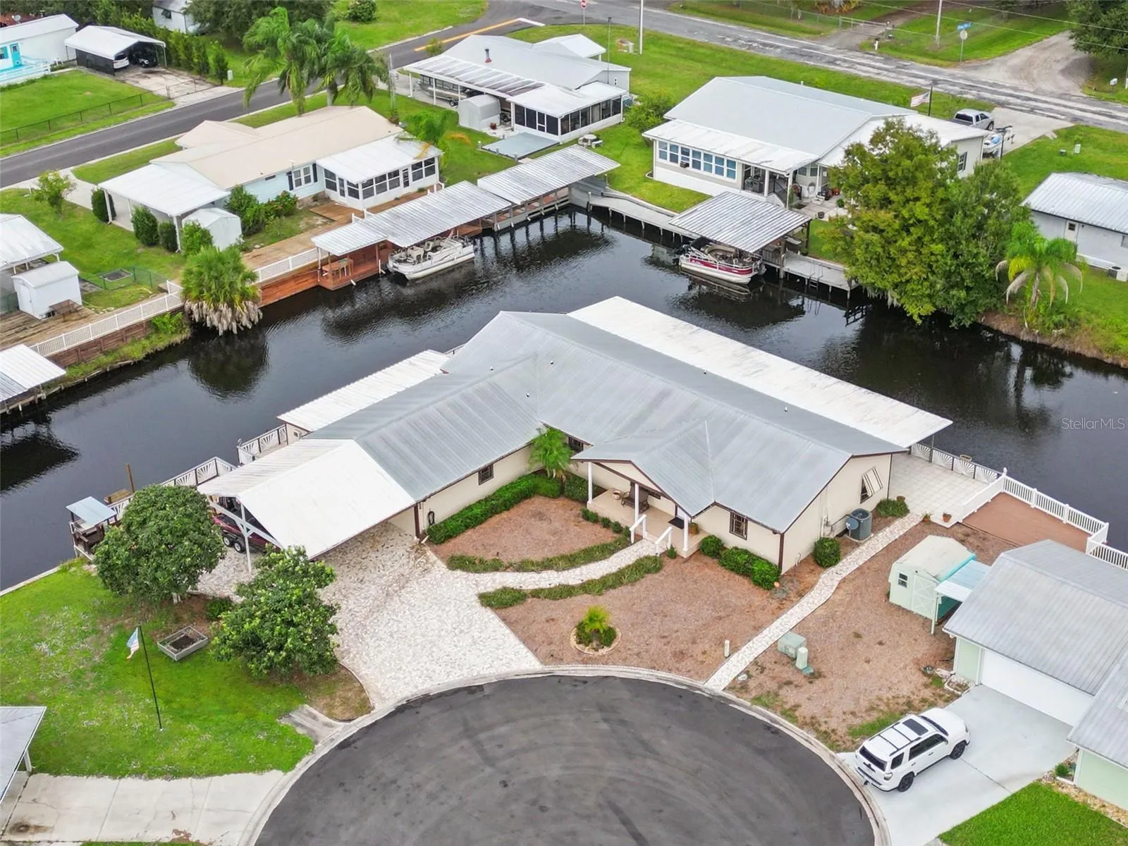 Aerial Waterfront Corner Lot: High-angle drone perspective showcasing the home's cul-de-sac location, metal roofing, extended carport, and extensive canal frontage