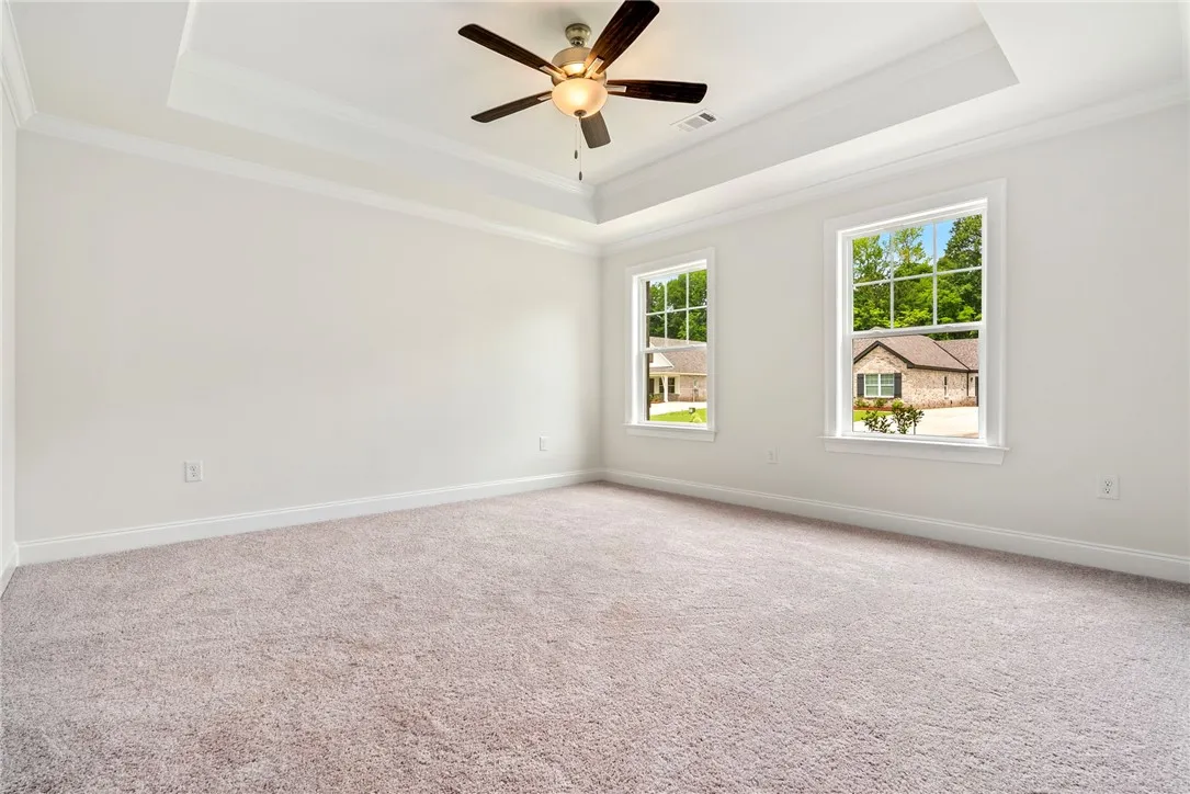 A spacious bedroom showcases a tray ceiling with a fan and twin sunlit windows overlooking the neighborhood.