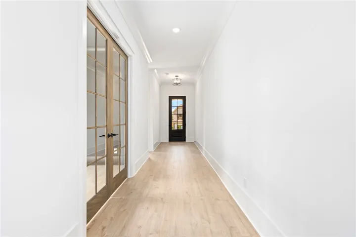 A long, bright corridor with light hardwood flooring leads to a dark paneled door with glass inserts.