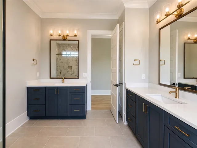 A well-lit primary bathroom features dual vanities with deep blue cabinetry and contemporary brass fixtures.