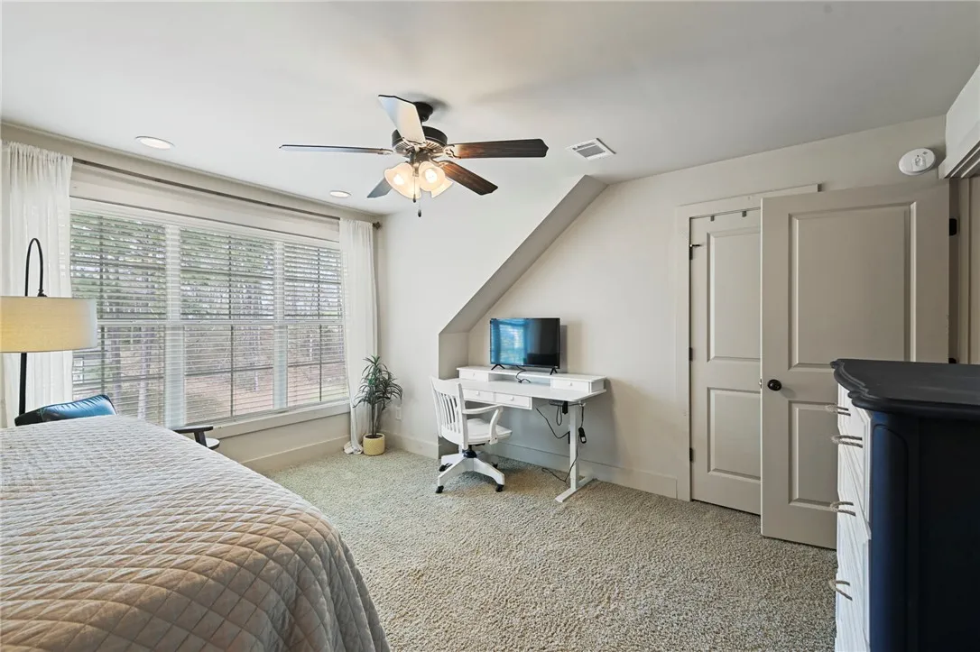 A well-lit bedroom with a ceiling fan, plush carpet, and ample natural light from a wide window.