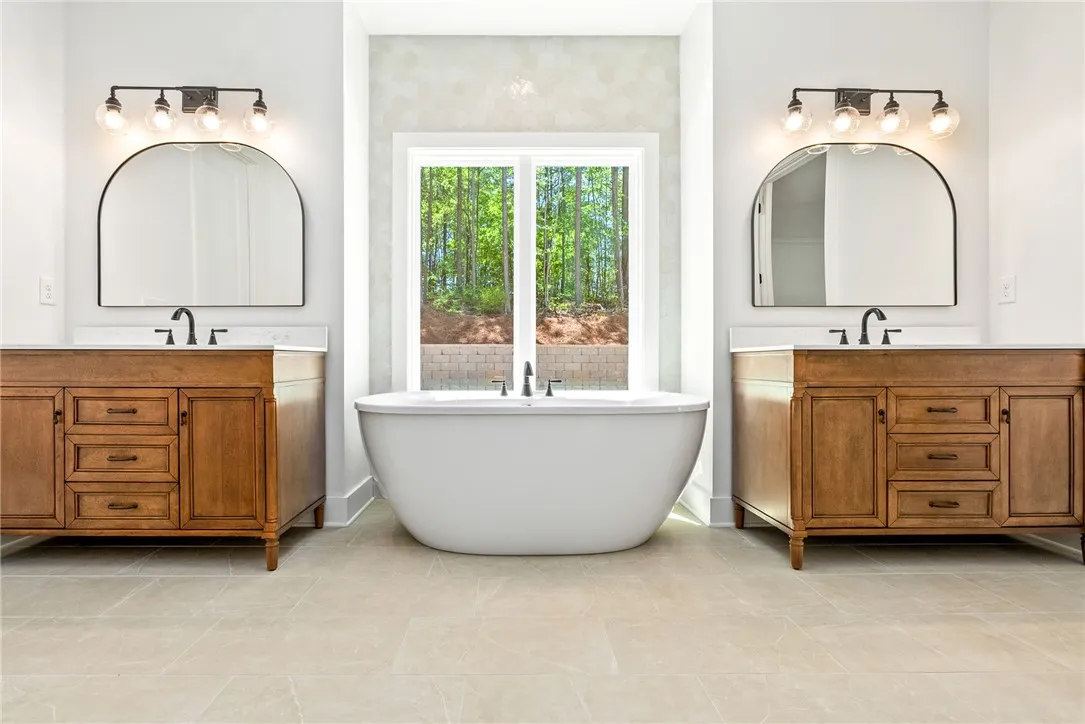 This primary bath features a soaking tub, dual furniture-influenced vanities, and a serene view.