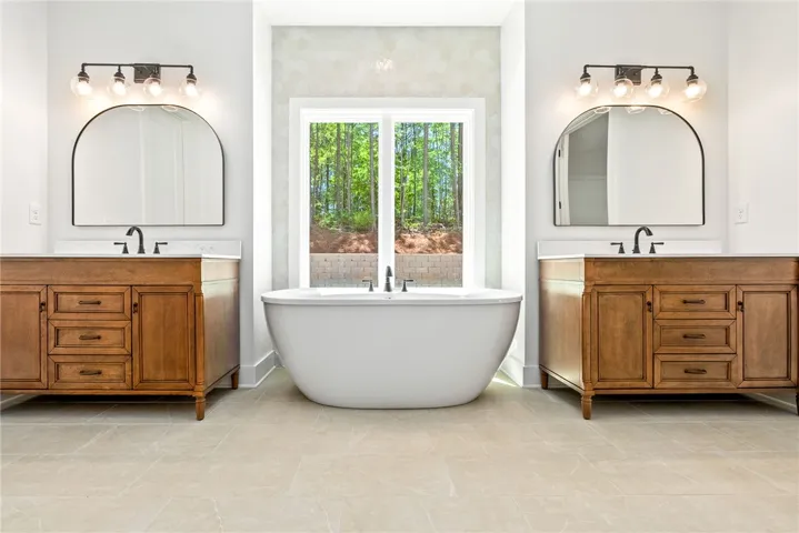 This primary bath features a soaking tub, dual furniture-influenced vanities, and a serene view.