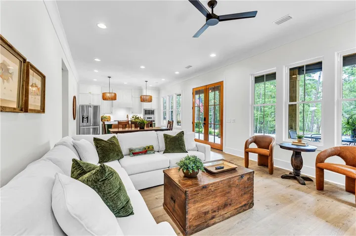 An airy living space with tall windows and light flooring connects seamlessly to a modern kitchen.