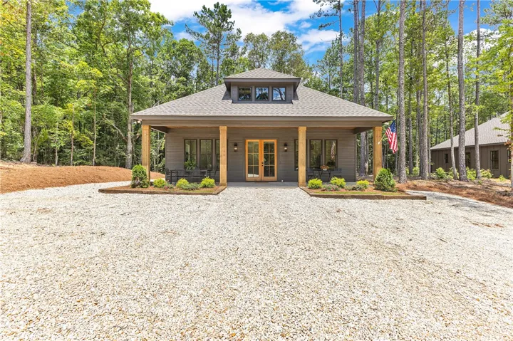 A welcoming front porch, shaded by towering trees, features natural wood columns and a gravel driveway.