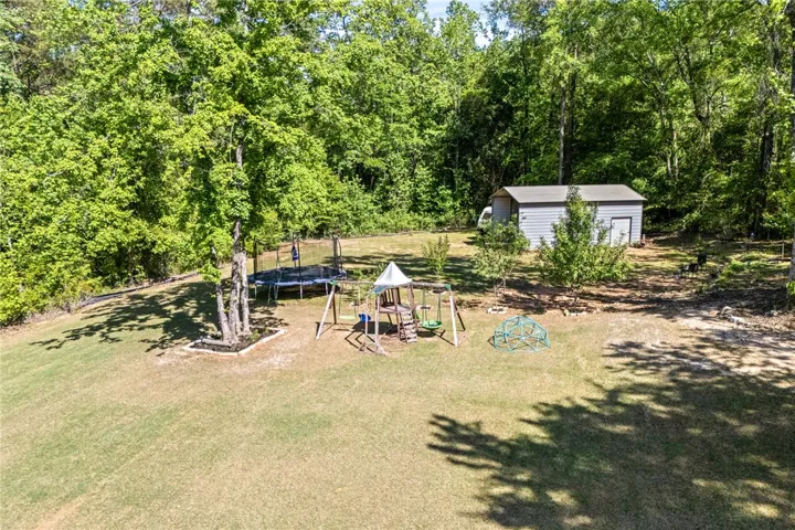 Expansive green grounds featuring a swing set, trampoline, and utility shed, enveloped by a lush tree line.