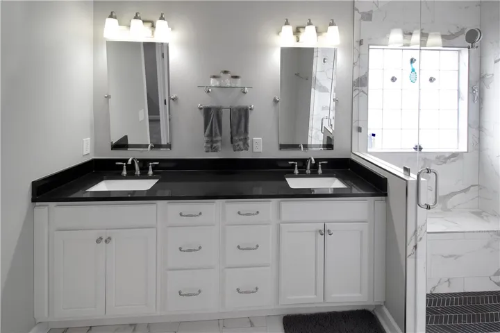 This spacious bathroom offers dual vanities with dark countertops and a bright, tiled shower.