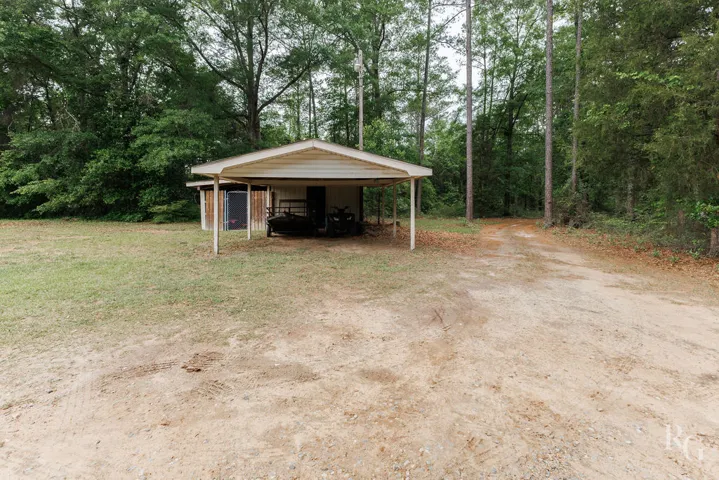 This property features a practical carport, ample parking, and convenient storage, all surrounded by lush trees.