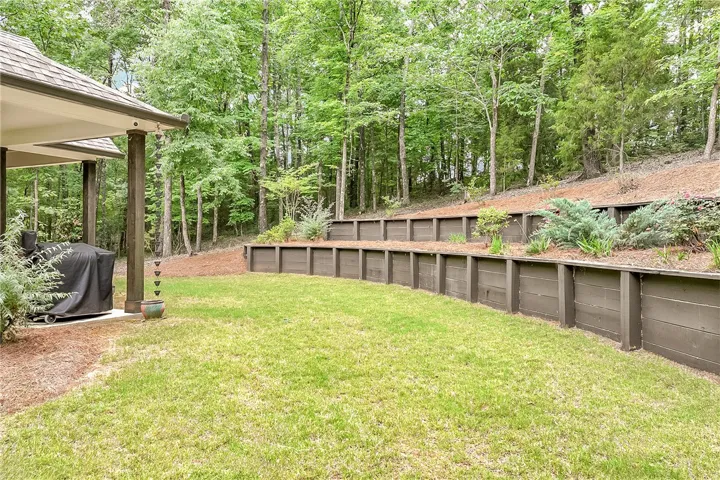 A well-maintained lawn and shaded patio area extend into a natural wooded setting, featuring terraced landscaping.