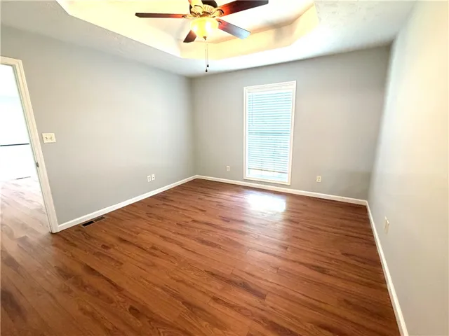 This primary bedroom showcases rich wood flooring and a distinctive tray ceiling with a fan.