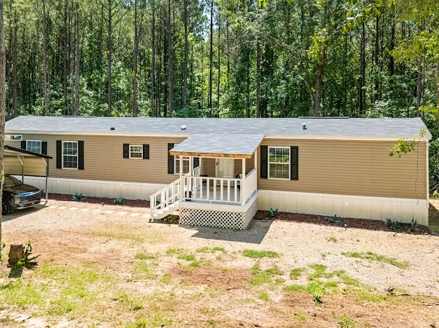A well-maintained manufactured home nestled among a lush tree line, featuring a welcoming covered porch and practical carport.