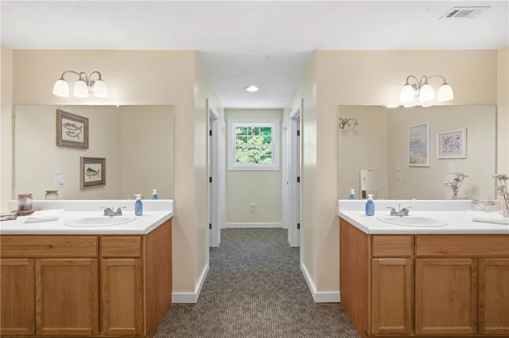 This functional bathroom features two separate vanities with expansive mirrors and a windowed hallway leading to additional private spaces.
