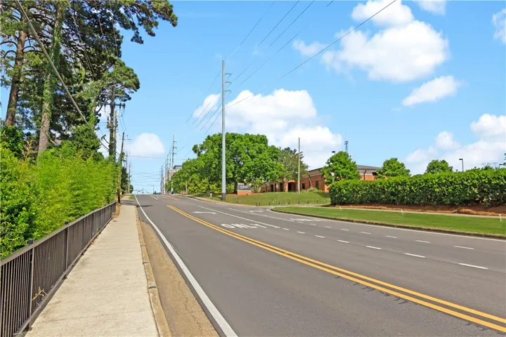 A wide, paved road with sidewalks, flanked by verdant trees and manicured bushes under a bright sky.