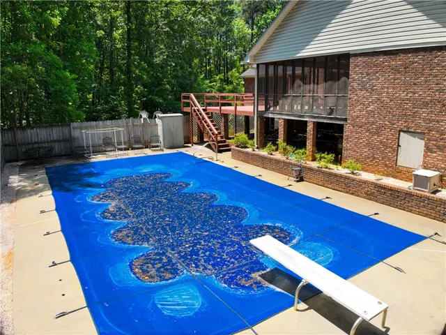 A spacious outdoor pool area, bordered by a fenced yard and a classic brick-sided residence with a screened porch.