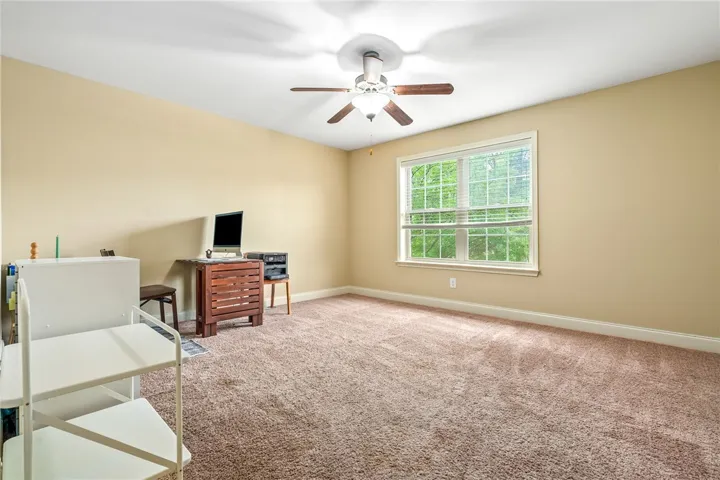 An inviting room features plush carpeting and abundant natural light from a broad window, centered by a ceiling fan.