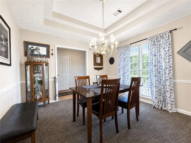 This formal dining space features tray ceilings, carpeted flooring, and a large window with patterned drapes.