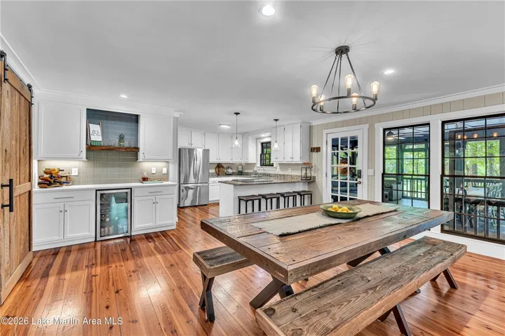 A well-lit kitchen with natural wood flooring and a large, rustic dining table flows into a bright screened porch.