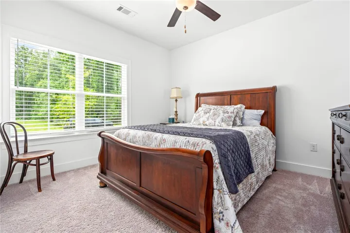 A well-lit bedroom with a ceiling fan, plush carpet, and large windows offering verdant views.