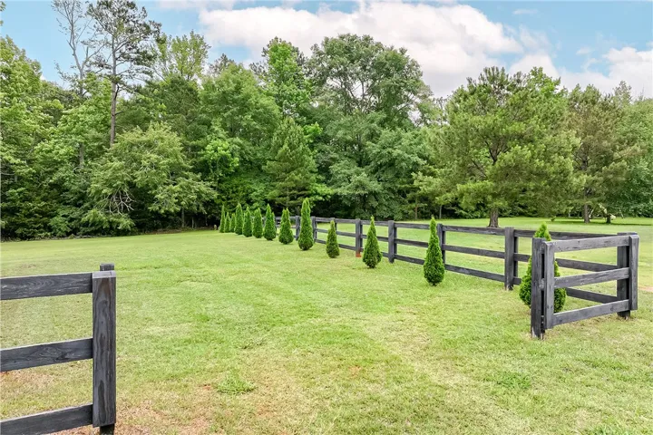 A dark-stained wooden fence defines the property line, backed by a lush tree line and punctuated by a row of small evergreens.