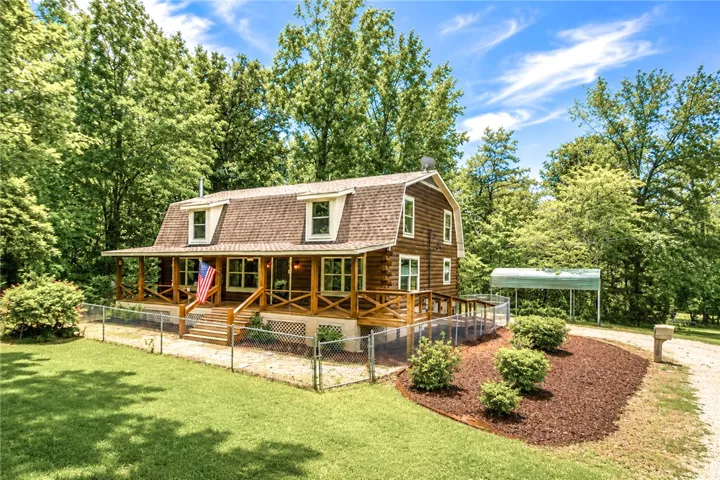 A charming log home with a welcoming covered porch, fenced yard, and lush landscaping, nestled among mature trees.