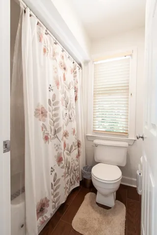 A well-lit full bathroom features dark hardwood floors, a bright window, and a shower with a floral curtain.