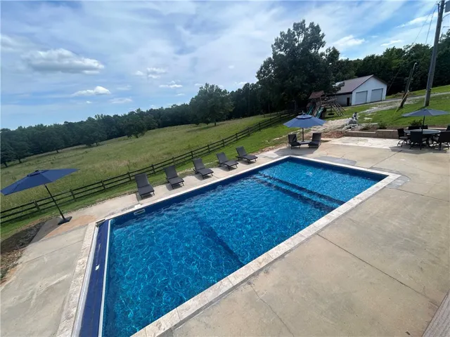 An inviting in-ground pool beckons with lounge seating and ample patio space, set against a tranquil, open field.