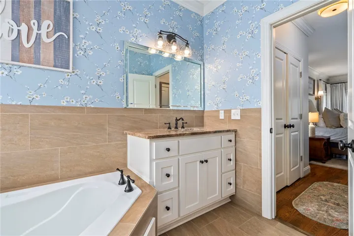 A well-lit primary bathroom features a corner tub, a granite-topped vanity, and delicate floral wallpaper.